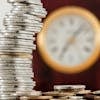 A close-up image of stacked coins with a blurred clock, symbolizing time and money relationship.