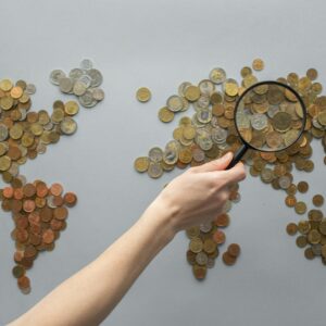 Top view of crop unrecognizable traveler with magnifying glass standing over world map made of various coins on gray background