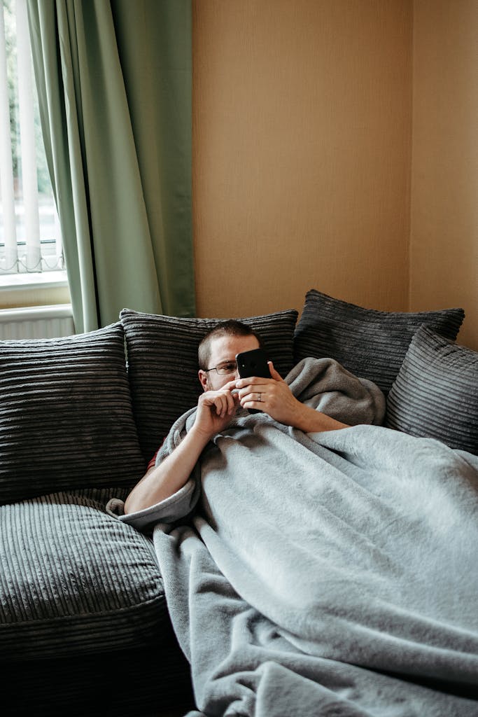 Casual indoor scene of a man lying on sofa, wrapped in a blanket, using smartphone.