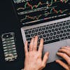 Hands typing on a laptop with cryptocurrency charts and smartphone display in view, focusing on digital finance.