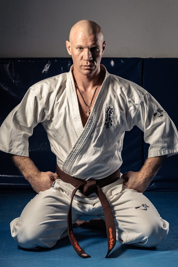 Karate practitioner kneeling in a dojo wearing a brown belt and white gi, demonstrating focus.